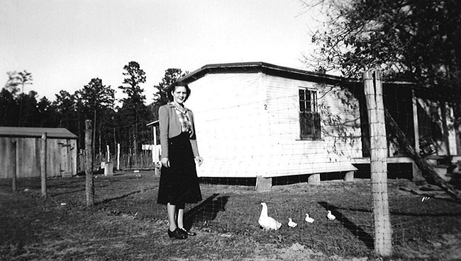 Young white woman in dress and chickens standing outside house with fence and outbuilding