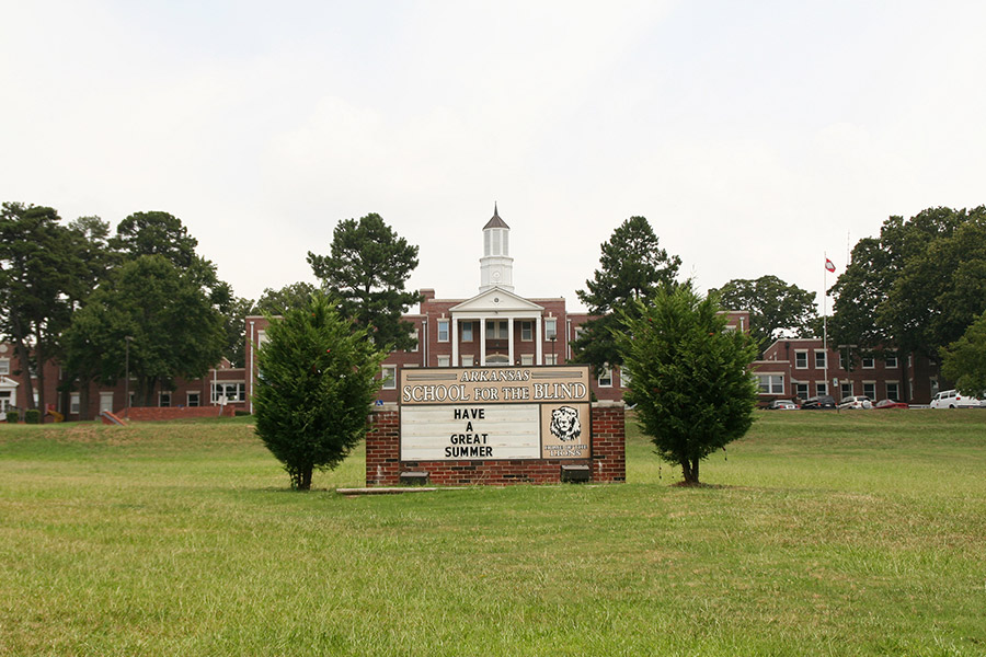 Multistory brick building with cupola on main section's roof and wings with trees and sign in front yard