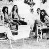 Five white men with long hair on lawn furniture below tree, accompanied by two white women
