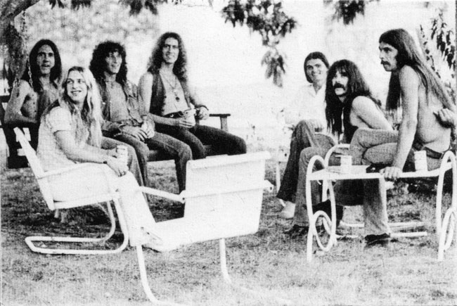 Five white men with long hair on lawn furniture below tree, accompanied by two white women
