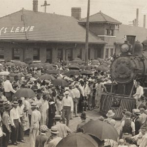Large crowd gathered around steam train at depot building with words saying "one hundred and twenty boys leave" written on photo