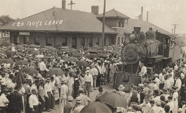 Large crowd gathered around steam train at depot building with words saying "one hundred and twenty boys leave" written on photo