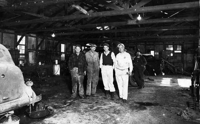 men posing in garage with cars in background and foreground