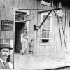 white man and little girl stand in entrance of wooden building. photo inset of white man's face in front of sign "Waiting Room for White Passengers"