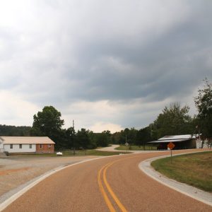 Single-story church building with steeple on left side of curved two-lane road across from other building