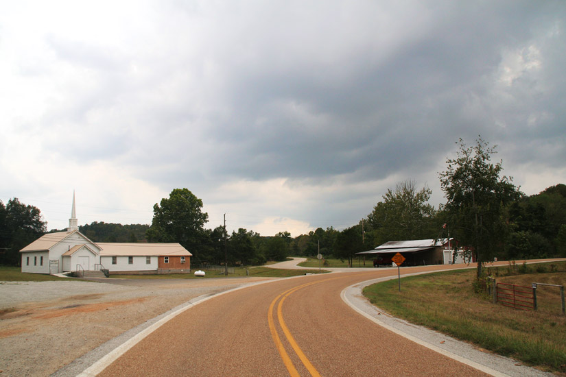 Single-story church building with steeple on left side of curved two-lane road across from other building