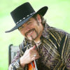 white man with anchor style goatee and mustache  in black and gold shirt and black cowboy hat holding guitar and smiling