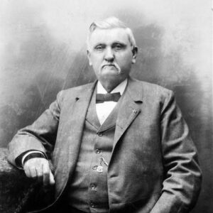 Portrait of a stern older white man seated on a wicker chair wearing a three-piece suit and a Confederate lapel pin