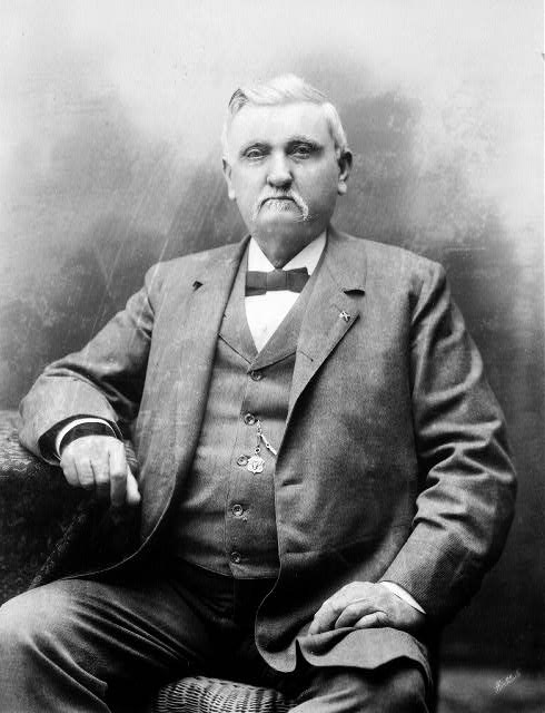 Portrait of a stern older white man seated on a wicker chair wearing a three-piece suit and a Confederate lapel pin