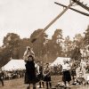 White man in kilt tossing a telephone pole as white spectators watch with tents in the background