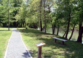 Gravel sidewalk in park lined with text displays on posts surrounded by trees and along a river