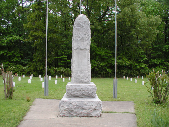 rough hewn stone monument with names engraved  standing before three flagpoles amid larger cemetery