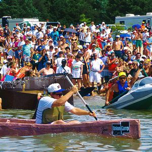 White men racing cardboard boats with crowd