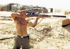 Shirtless white man aiming a sniper rifle with silencer