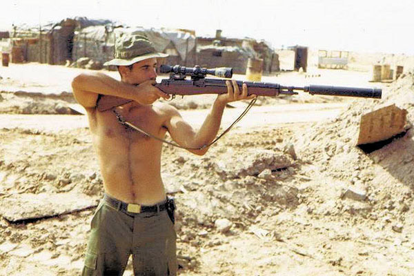Shirtless white man aiming a sniper rifle with silencer