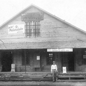 White man standing outside A-frame post office and general store building with covered porch