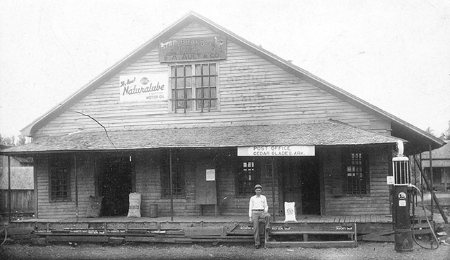 White man standing outside A-frame post office and general store building with covered porch