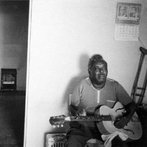 Interior two black men one seated with guitar one standing hands in pockets smiling