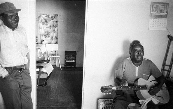 Interior two black men one seated with guitar one standing hands in pockets smiling