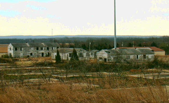 Run down single and multistory buildings and large flag pole with overgrown concrete foundations in the foreground