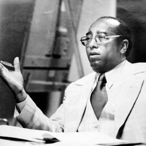 Seated black man wearing glasses and a suit and tie gestures with a pen in his hand in front of notebooks on a desk