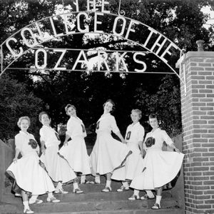 Young white women in cheerleader uniforms at entrance arch with words "College of the Ozarks"