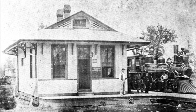 Small train station with wrap around awning pitched roof white people standing by locomotive