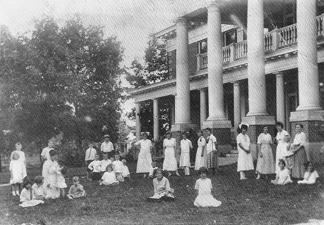 Group of women and children outside two-story house with four columns and balcony