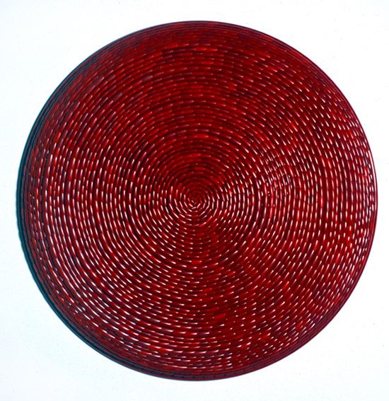 Low relief circular sculpture consisting of spiraling red fingernail tips resembling woven mat