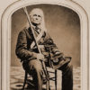 Old man with long hair and gun sitting in military uniform
