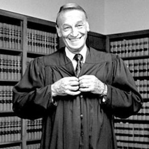 White man standing and smiling in his judge's robes with bookshelves behind him