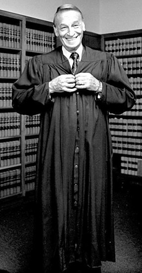 White man standing and smiling in his judge's robes with bookshelves behind him