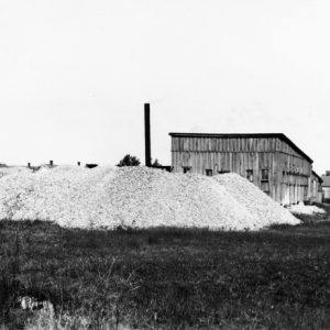 large pile of material in field outside wood-frame building with smoke stack