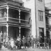 Group photo of white men and women in yard and on balcony outside multistory brick building
