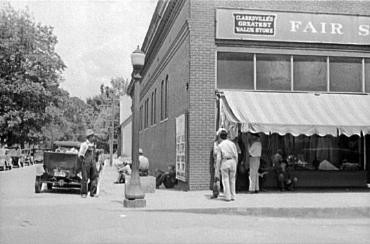 Street corner with men seated near cars and standing outside "Clarksville's greatest value store"