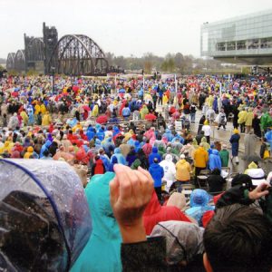 Large crowd gathering in brightly colored rain jackets by glass windowed modern building and steel bridge
