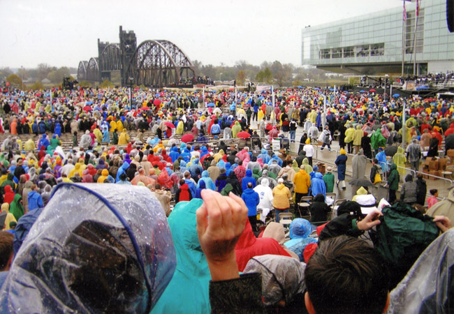 Large crowd gathering in brightly colored rain jackets by glass windowed modern building and steel bridge