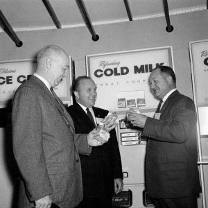 three white men in suits drink boxes of milk before vending machine labeled "Refreshing Cold Milk"