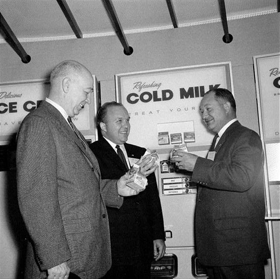 three white men in suits drink boxes of milk before vending machine labeled "Refreshing Cold Milk"
