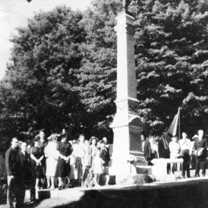 White people dressed formally standing below war monument, with large trees in background