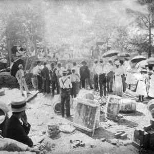 Crowd of men in hats and women with umbrellas gathering around brick foundation construction materials