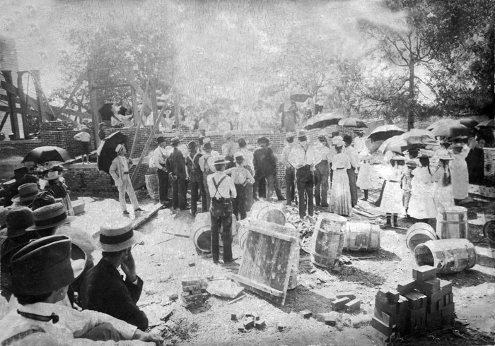 Crowd of men in hats and women with umbrellas gathering around brick foundation construction materials