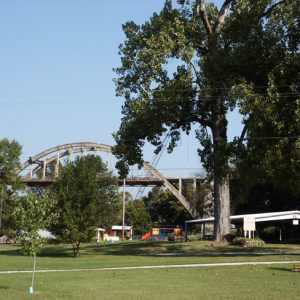 Park lawn including sidewalk large and small trees playground and large bridge in distance