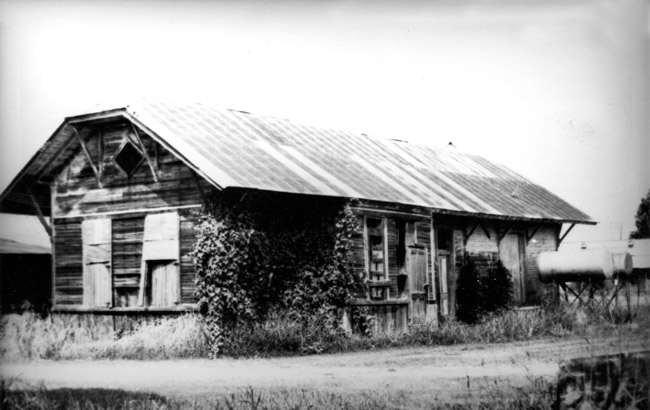 Wood frame building with metal roof boarded windows overgrown vines and water tanks by road