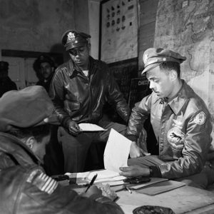 African American men in aviator jackets and hats around table looking at papers with maps on the walls