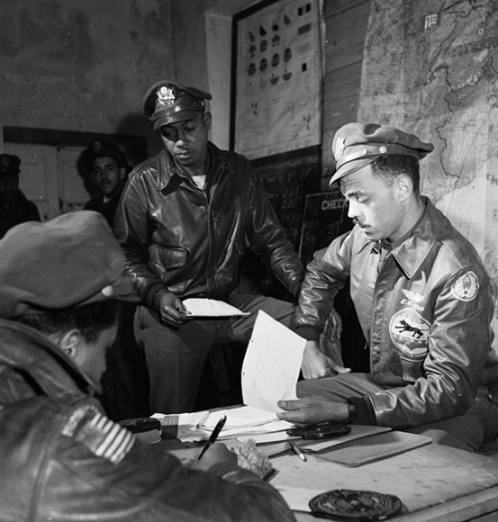 African American men in aviator jackets and hats around table looking at papers with maps on the walls