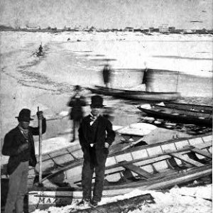 Men in dark suits and hats and rowboats on snowy river bank with other boaters pushing down icy rivulet