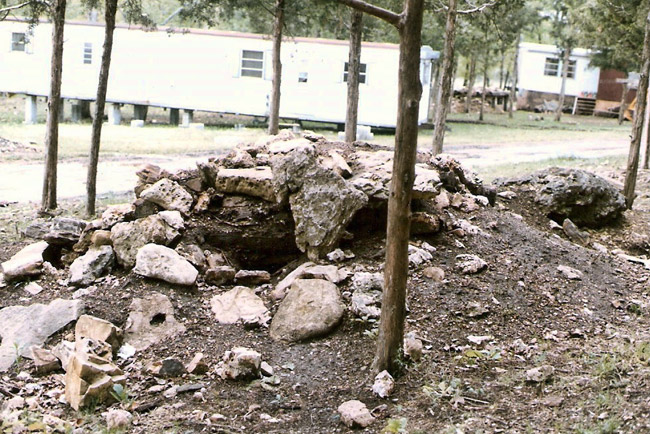 Underground bunker covered in dirt and rocks in foreground, trailer homes on cinder blocks in background