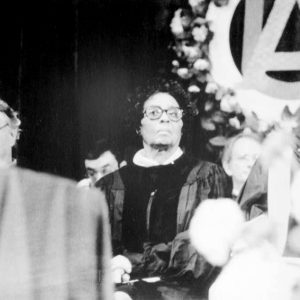Black woman in glasses and academic robes seated on stage amid white men