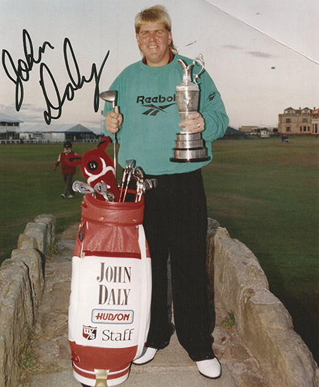 White man in green shirt holding a trophy in his left hand and golf club in his right with golf bag signed "John Daly"